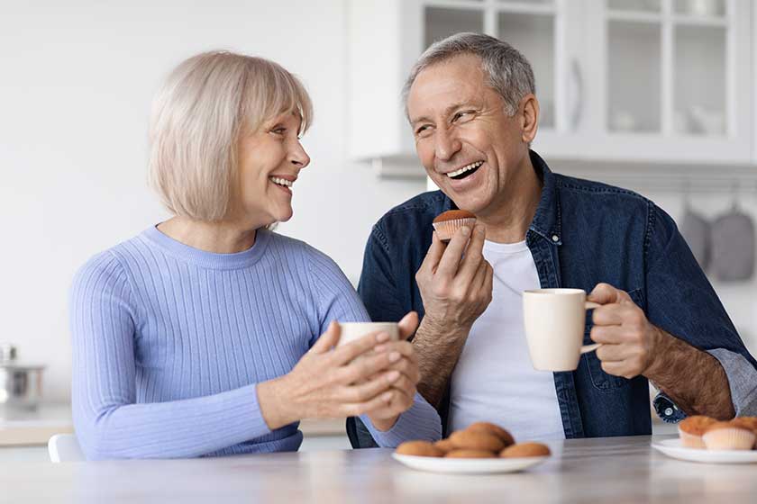 Happy beautiful elderly spouses drinking coffee with fresh pastry at home, sitting at kitchen table, holding mugs, having conversation and smiling, enjoying time together, closeup